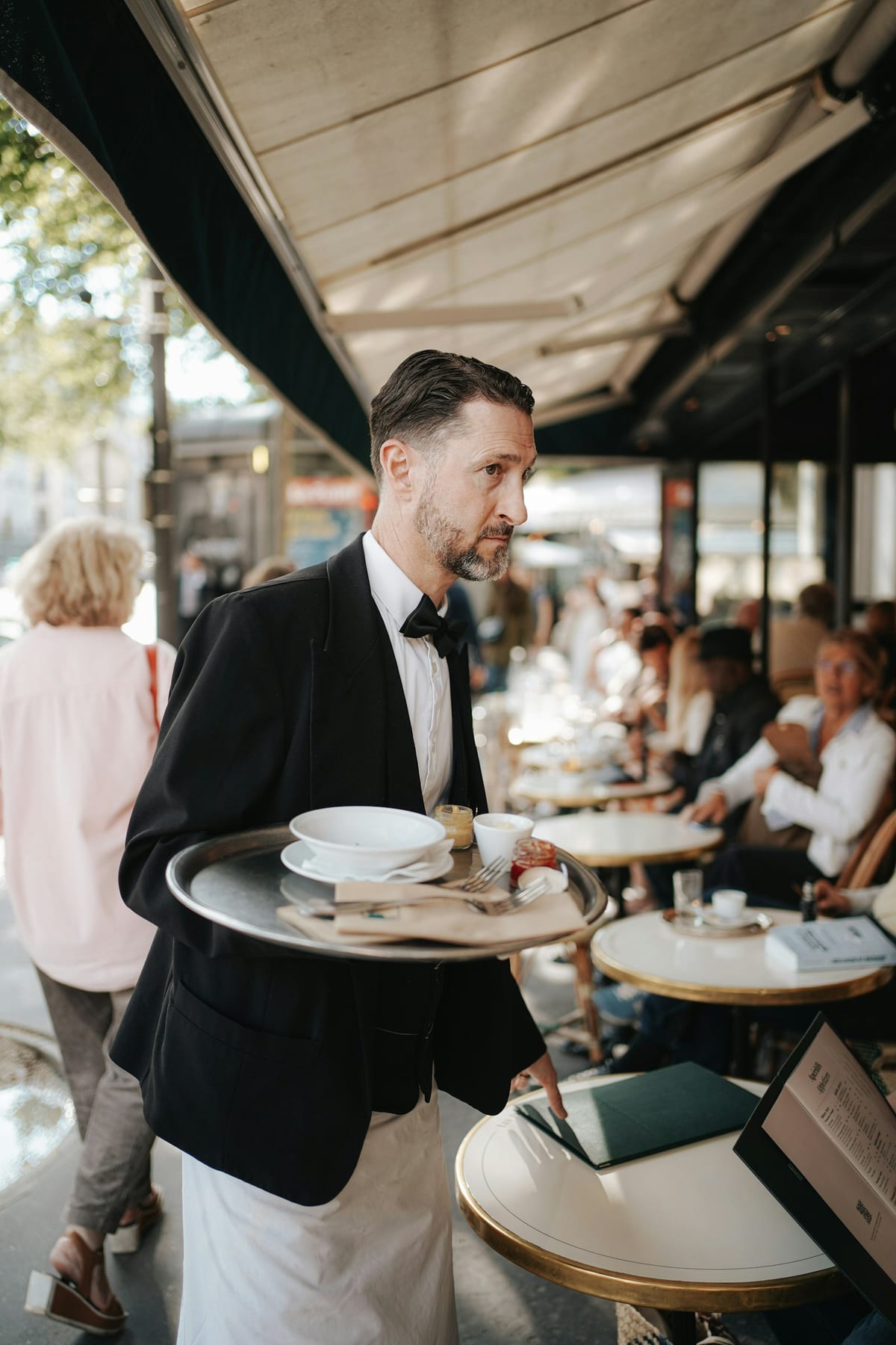 A waiter in a bow tie carrying a tray of crockery across a café terrace