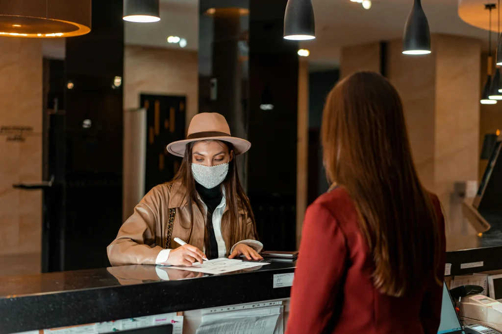 A front desk receptionist assisting a masked guest, highlighting customer service expertise as one of the key skills needed for hospitality.